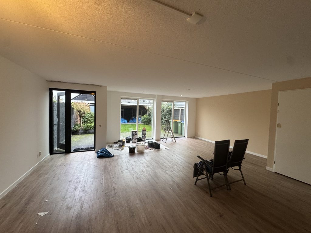 Lege woonkamer van een eengezinswoning met tuindeuren, een houten vloer en verfemmers op folie